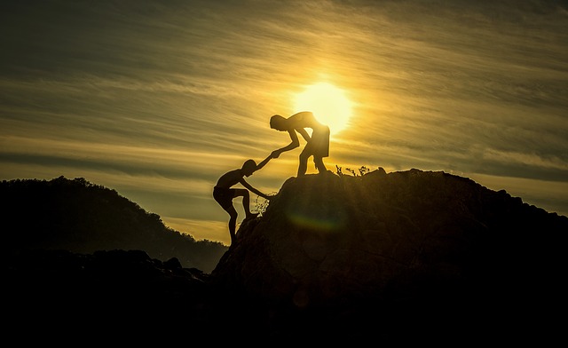 one man helping another climb to the peak of a mountain, while the sun sets in the background.