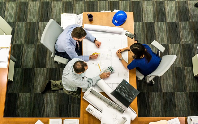 three business workers, hudled over a desk, working on large pieces of paper.